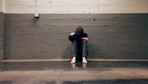 Young person sitting isolated against a wall hunched over with their head down.